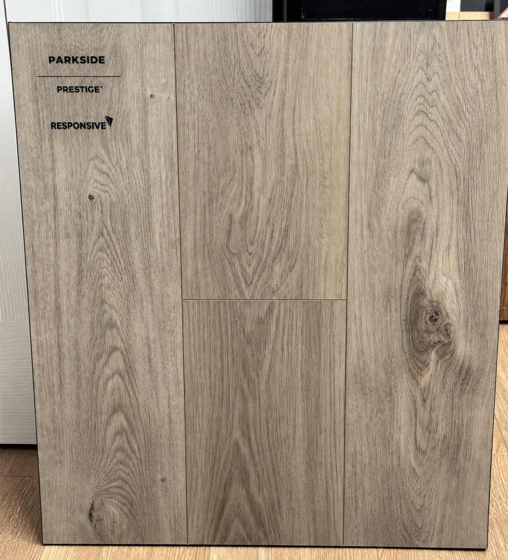 A large square sample of Parkside Prestige Responsive flooring shown in a light gray-brown woodgrain finish, standing upright indoors.