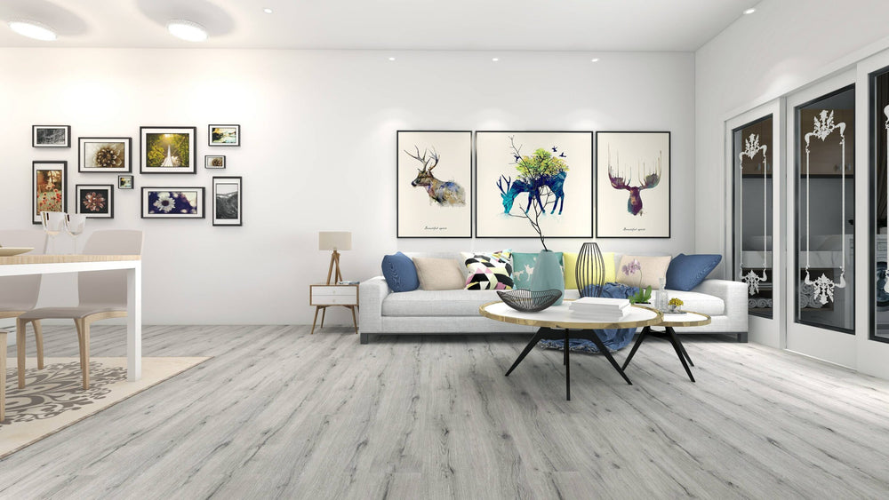 A modern living room with light gray wood-look flooring, a white sofa with blue and black accent pillows, a round coffee table, and framed wall art above the couch. The room is brightly lit with recessed ceiling lights and appears to be a 3D interior render.
