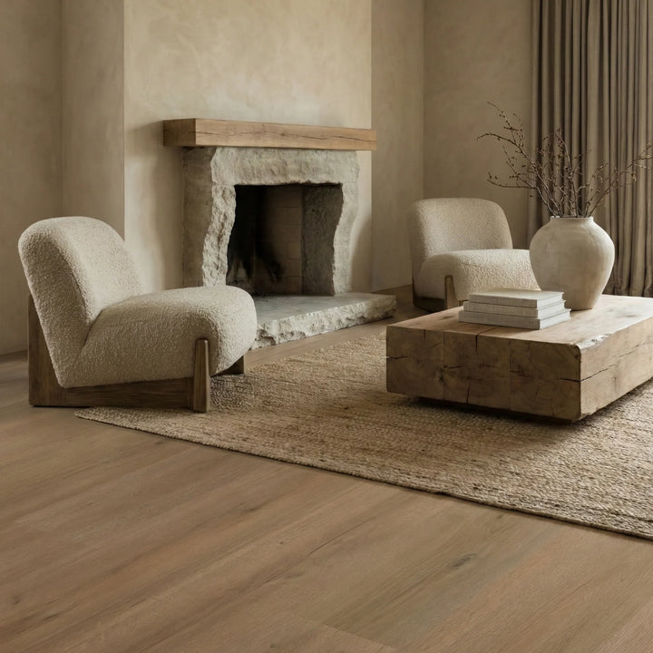 A styled living room scene with two cream boucle armchairs, a rustic wooden coffee table, a woven rug, a stone fireplace, and a ceramic vase on a light wood floor.