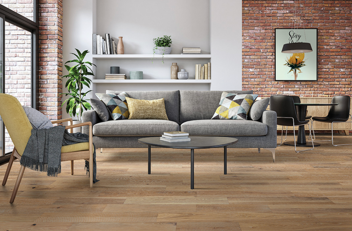 A modern living room scene featuring a gray fabric sofa with decorative pillows, a round black coffee table, a yellow armchair with a gray throw, a brick wall, and a framed art print.
