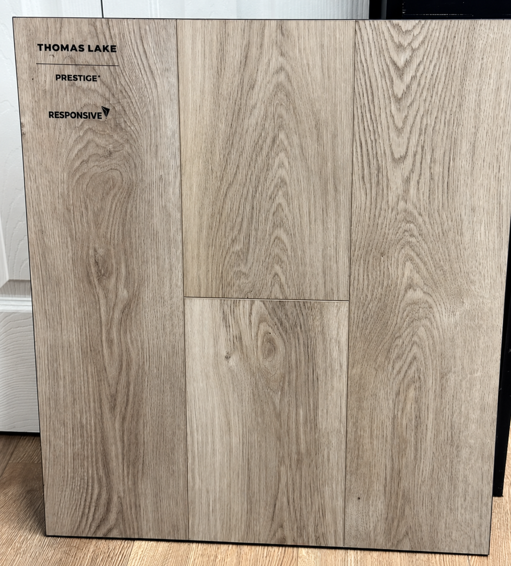 A large square wood-look flooring sample board standing upright indoors. The board shows three vertical plank sections with a light oak grain pattern and printed text reading THOMAS LAKE, PRESTIGE, and RESPONSIVE.