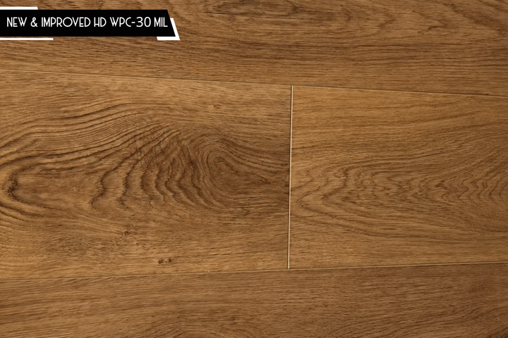 Oak wood-look flooring planks in a medium brown tone with visible grain patterns and a small black text banner in the top left corner reading 'NEW & IMPROVED HD WPC-30 MIL'.