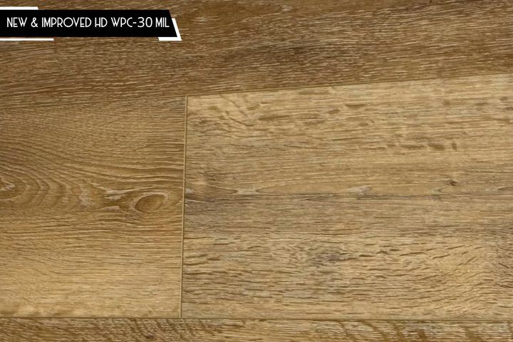 Close-up of a wood flooring sample showing grain texture and plank seams, with a small text label in the top-left corner reading 'NEW & IMPROVED HD WPC-30 MIL'.