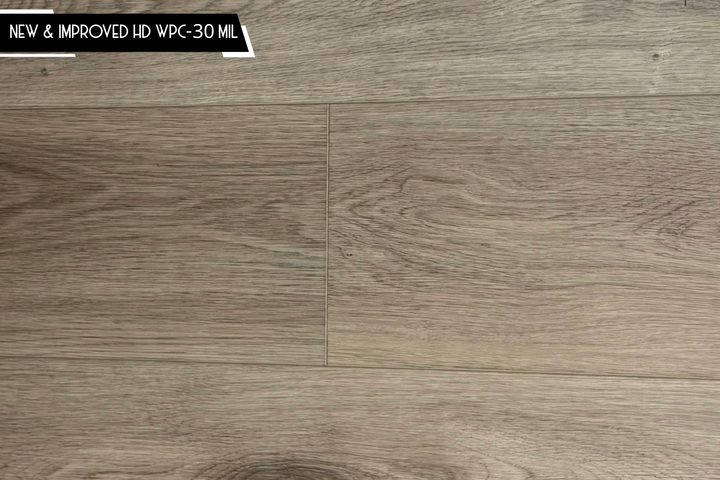 Close-up of wood-look flooring planks in a light taupe-brown tone with visible grain texture and a small black label in the top-left corner reading 'NEW & IMPROVED HD WPC-30 MIL'.